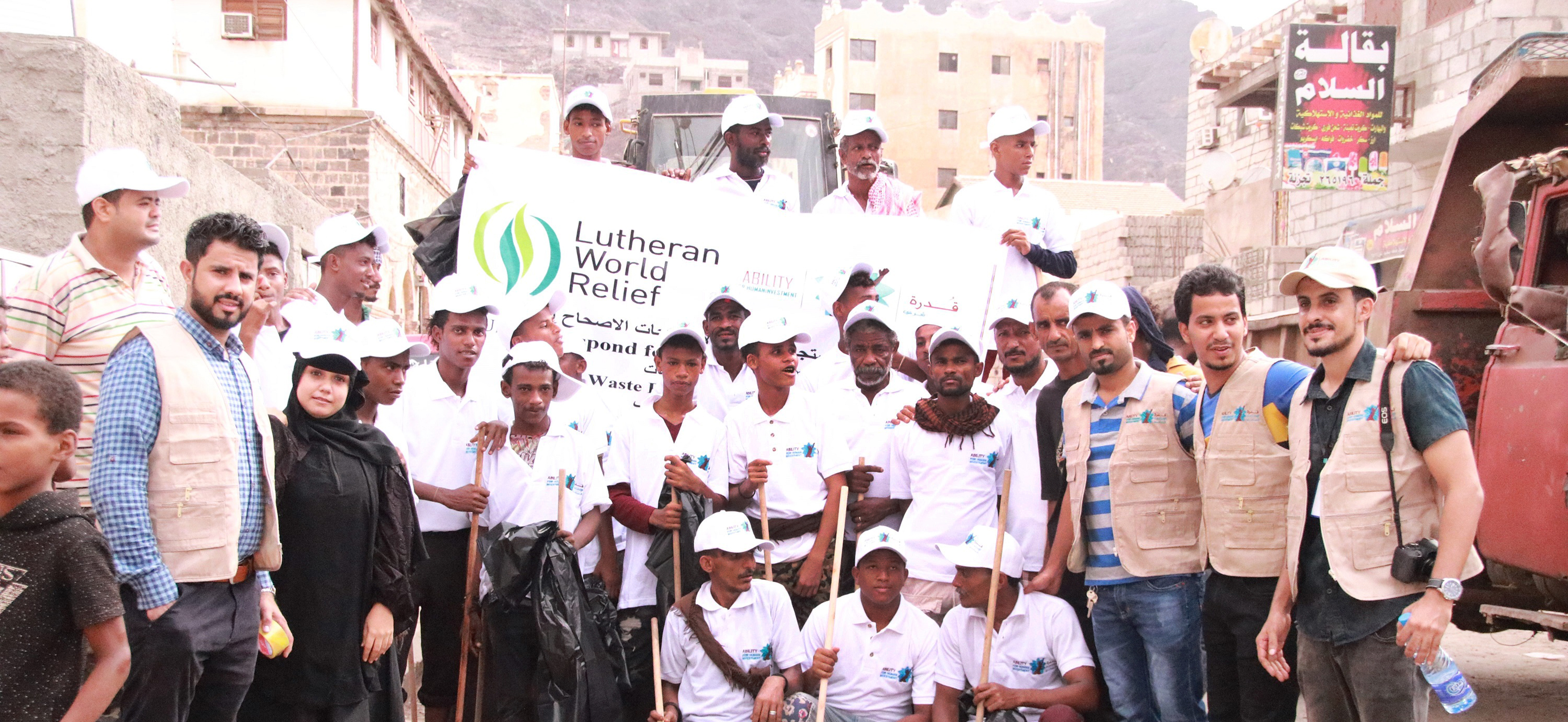 A group of LWR staff and local partner staff stand in the streets of Aden with a Lutheran World Relief sign after a cleanup campaign