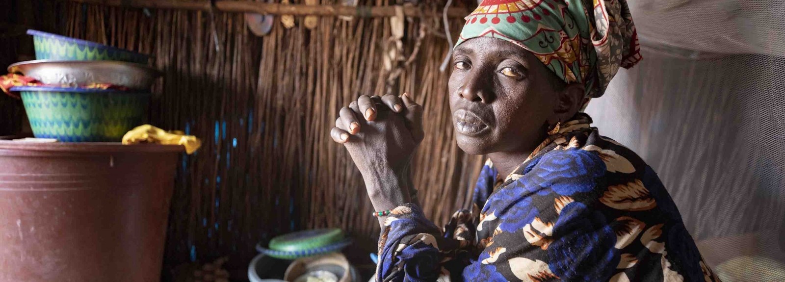 A woman with folded hands sits inside her home, which appears to be made of wooden sticks.made of sticks