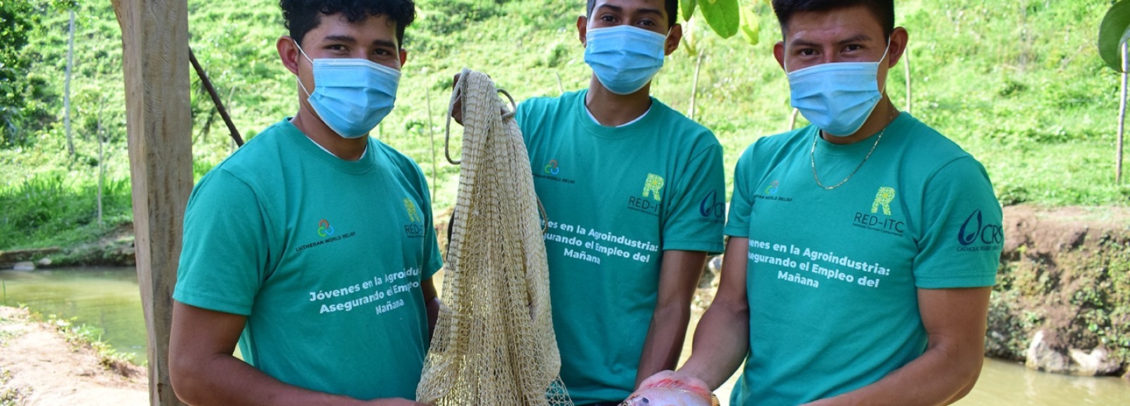 3 Honduran youth stand in front of a pond holding a fishing net and showing off a caught fish