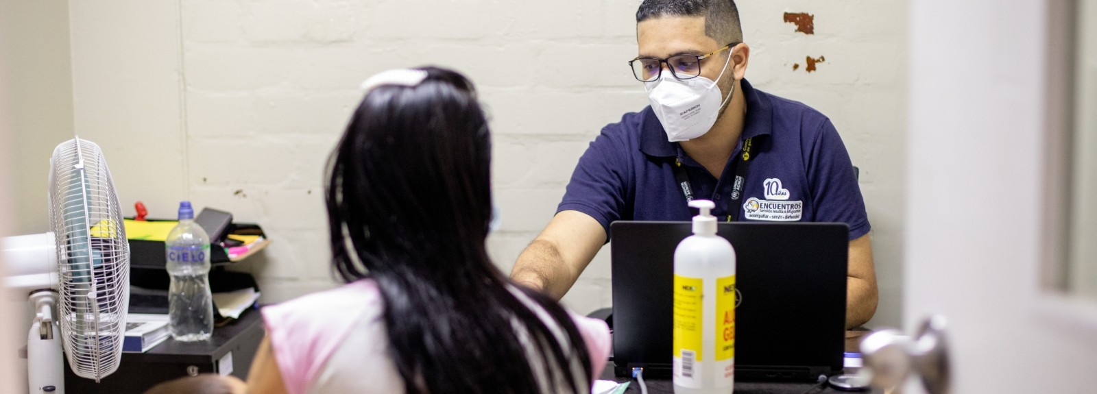 A lawyer in a mask sits behind his desk and his talking to a woman holding a baby
