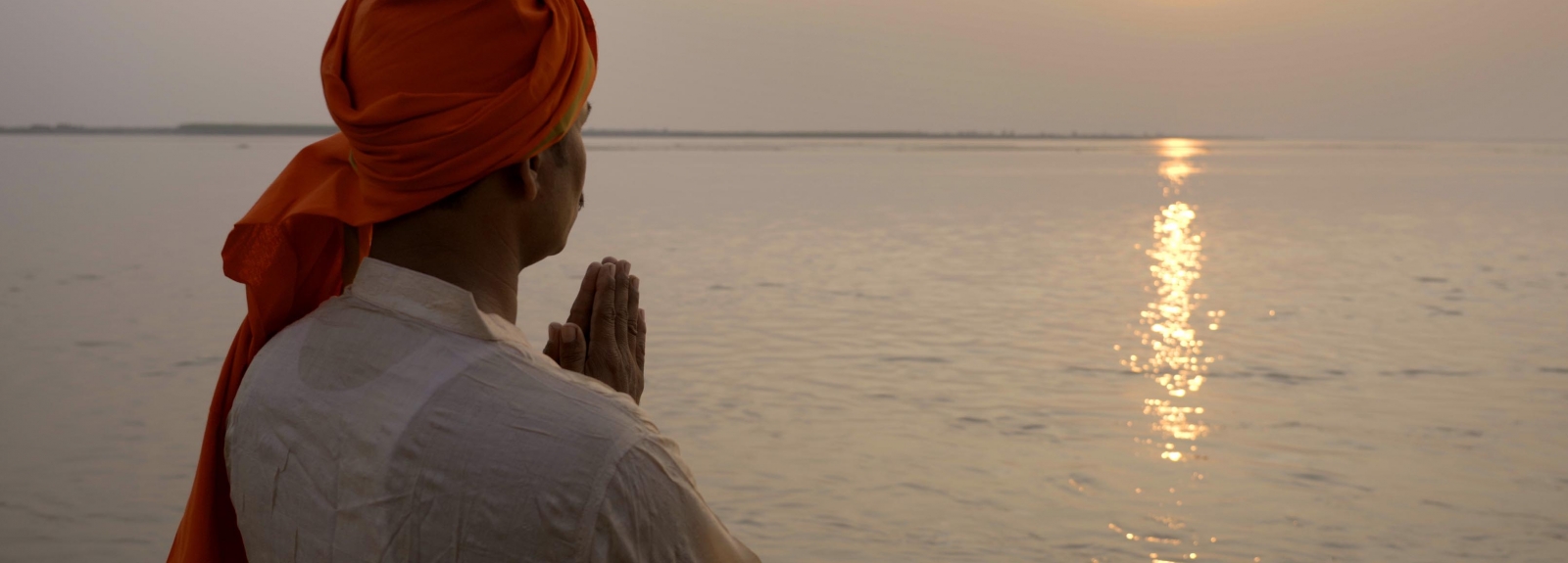 A man stands in front of a body of water with his hands folded in prayer.