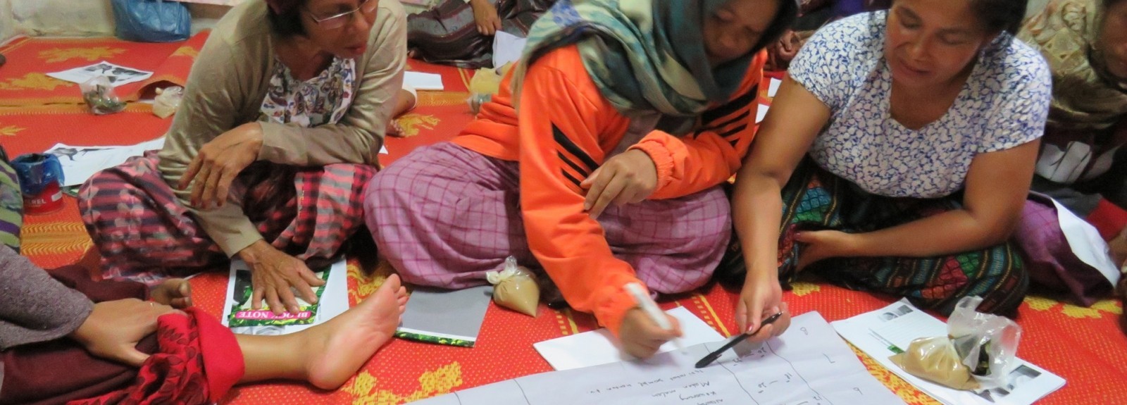 A group of women sit on a bright orange blanket writing on a large sheet of paper together