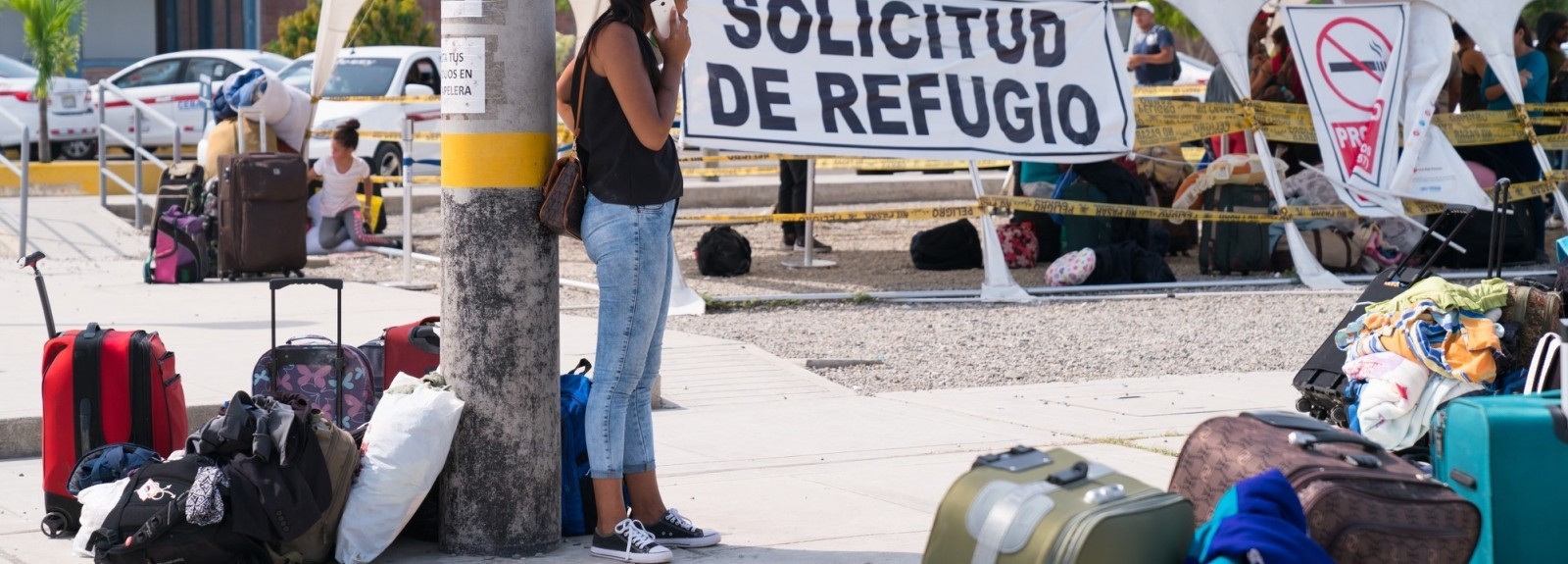 Woman stands next to a lamp post talking on her cell phone surrounded by luggage and belongings of fellow migrants at the border crossing