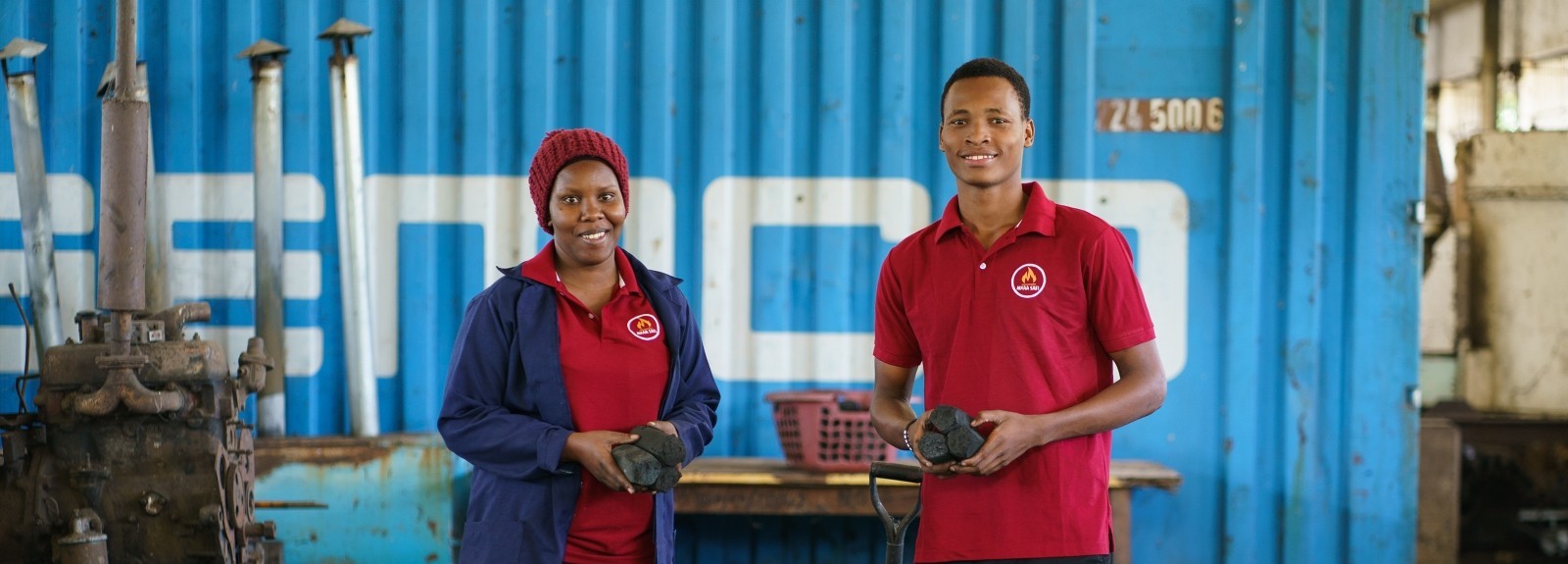 Two young Tanzanian entrepreneurs stand smiling in their workshop while holding charcoal