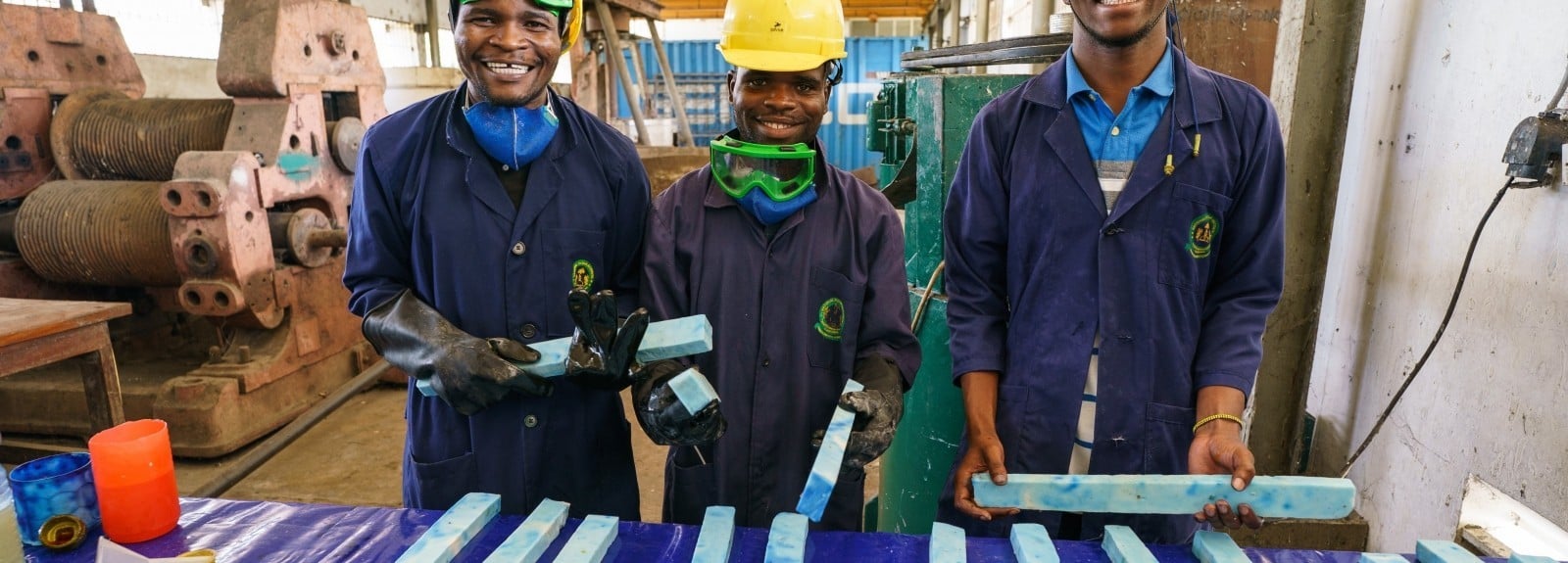 Three smiling young men wearing protective equipment in a warehouse hold columns of soap ready to be cut into bars 