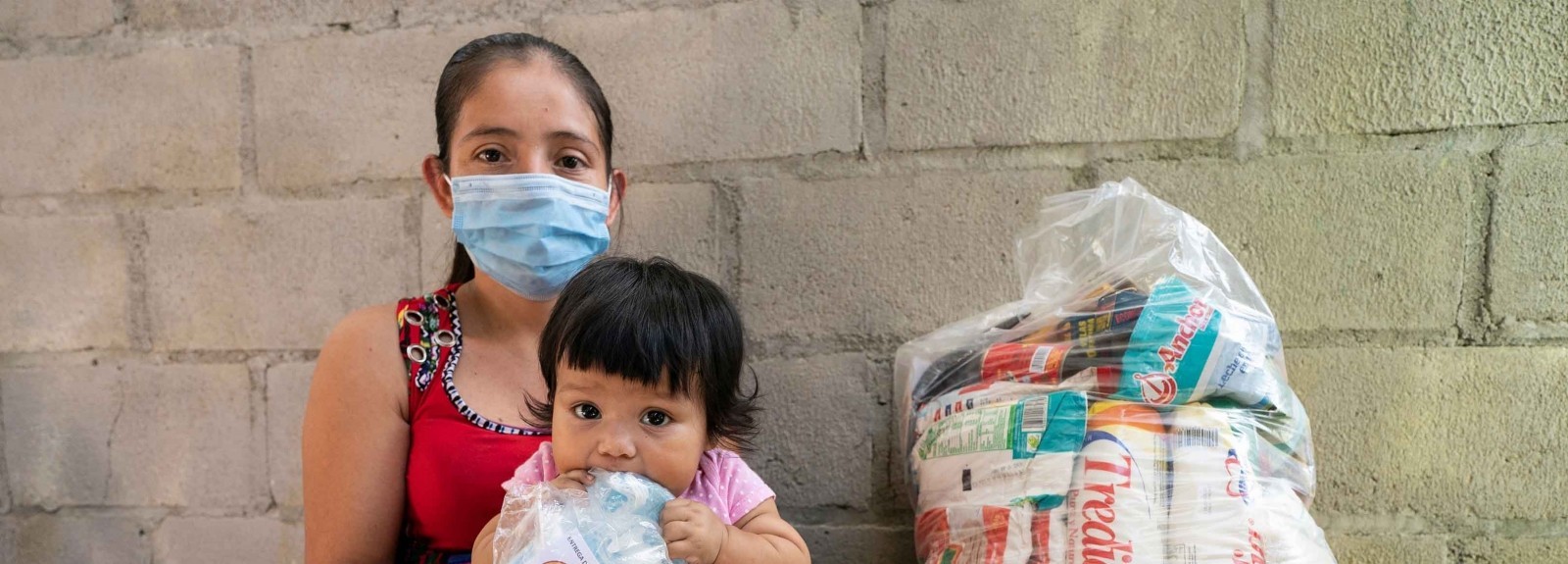 A woman, wearing a face covering, holds a young child, who is holding a bag. They are sitting next to a bag of necessities.