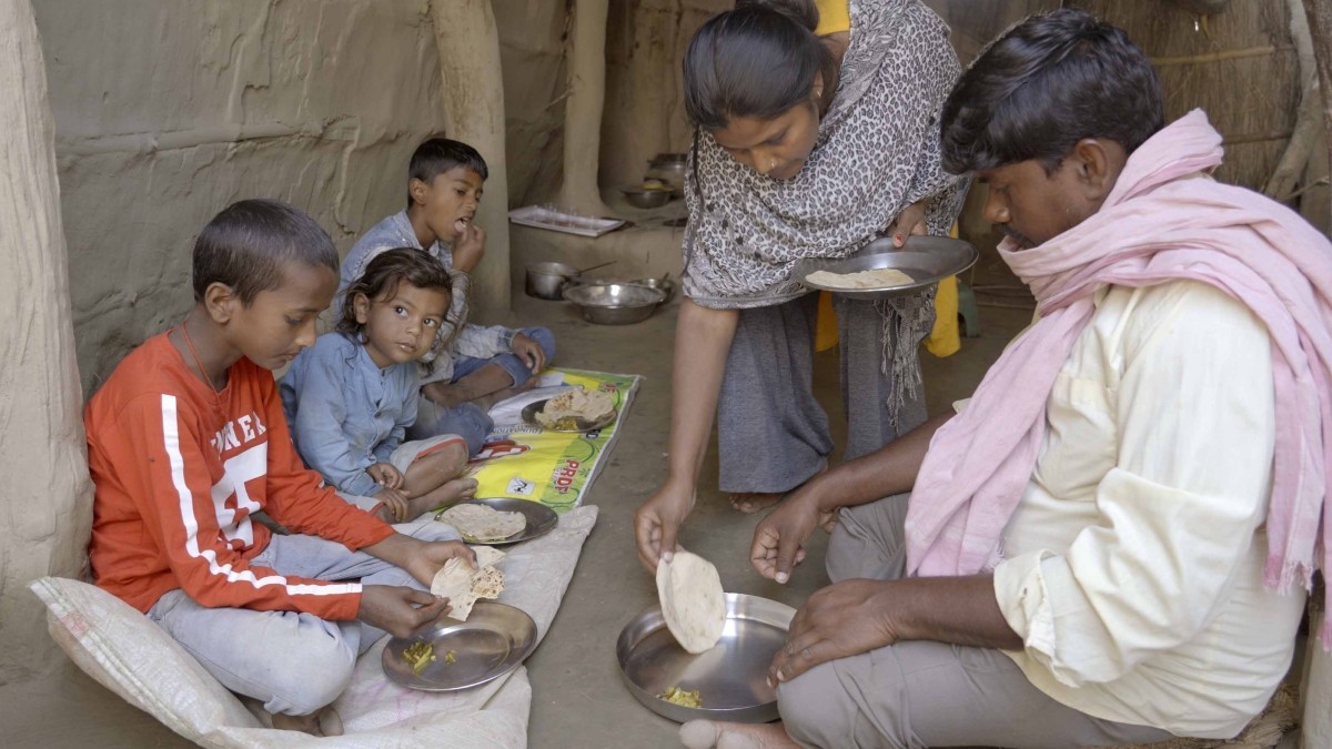 Three children, at left, are sitting on the floor with plates of food in front of them. A woman and a man are at right. The woman, who is standing, is leaning over to put food on the plate for the man, who is seated.