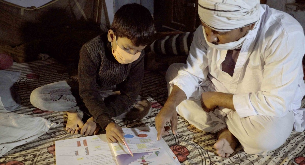 A child, left, and an adult, right, both wearing face coverings, look at a book together. The boy is holding a pen. They are sitting on the floor.