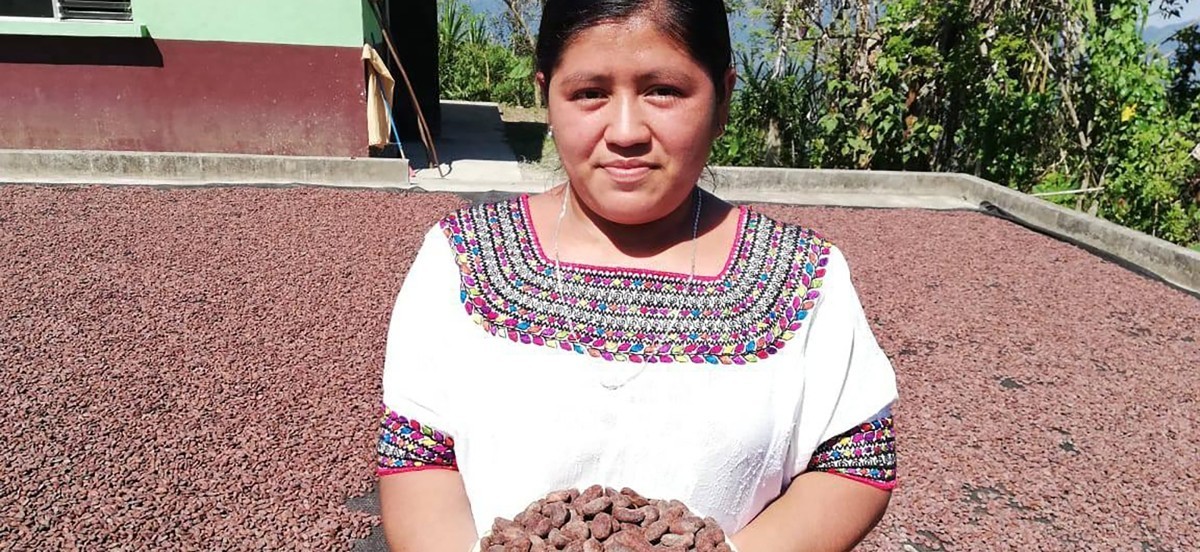A young woman holding dried cacao beans stands in front of a courtyard filled with cacao beans.