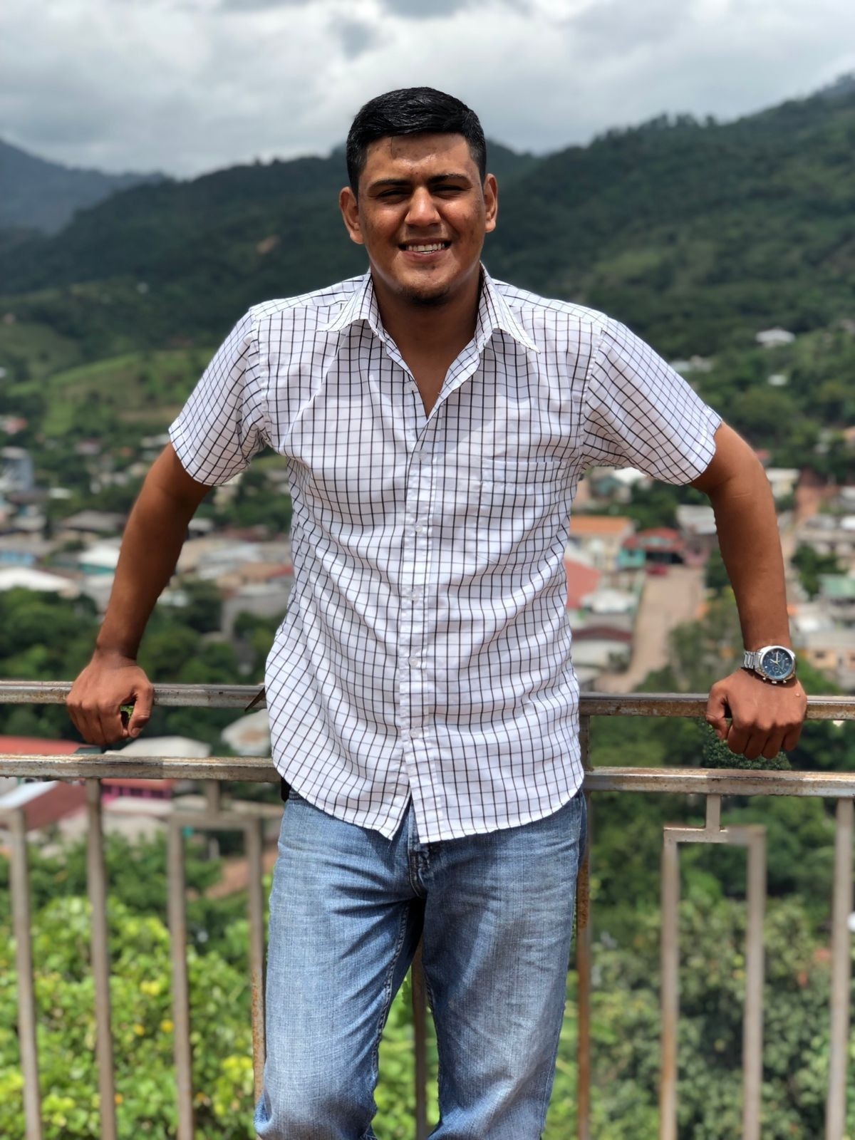 Young man in checkered white shirt stands smiling in front of a railing with green mountains in background