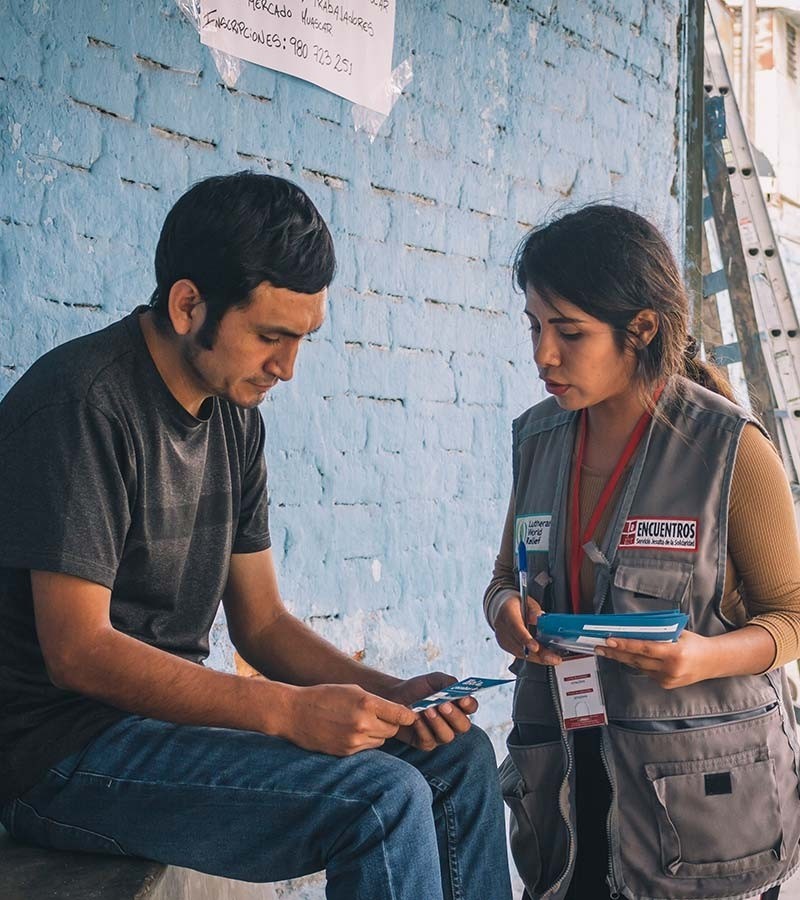 A VenInformado staff member in her cargo vest uniform shares information about the program with a man sitting in front of a blue wall