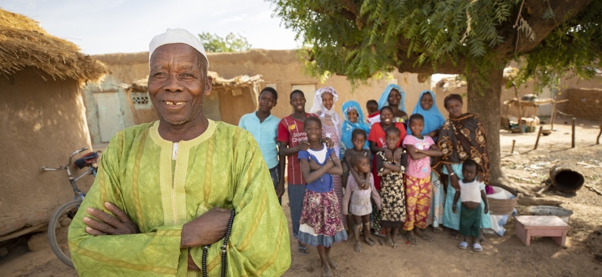 A 75-year-old Malian farmer in green clothes stands smiling at the front of the frame with his arms crossed while 14 members of his family stand in the background behind me