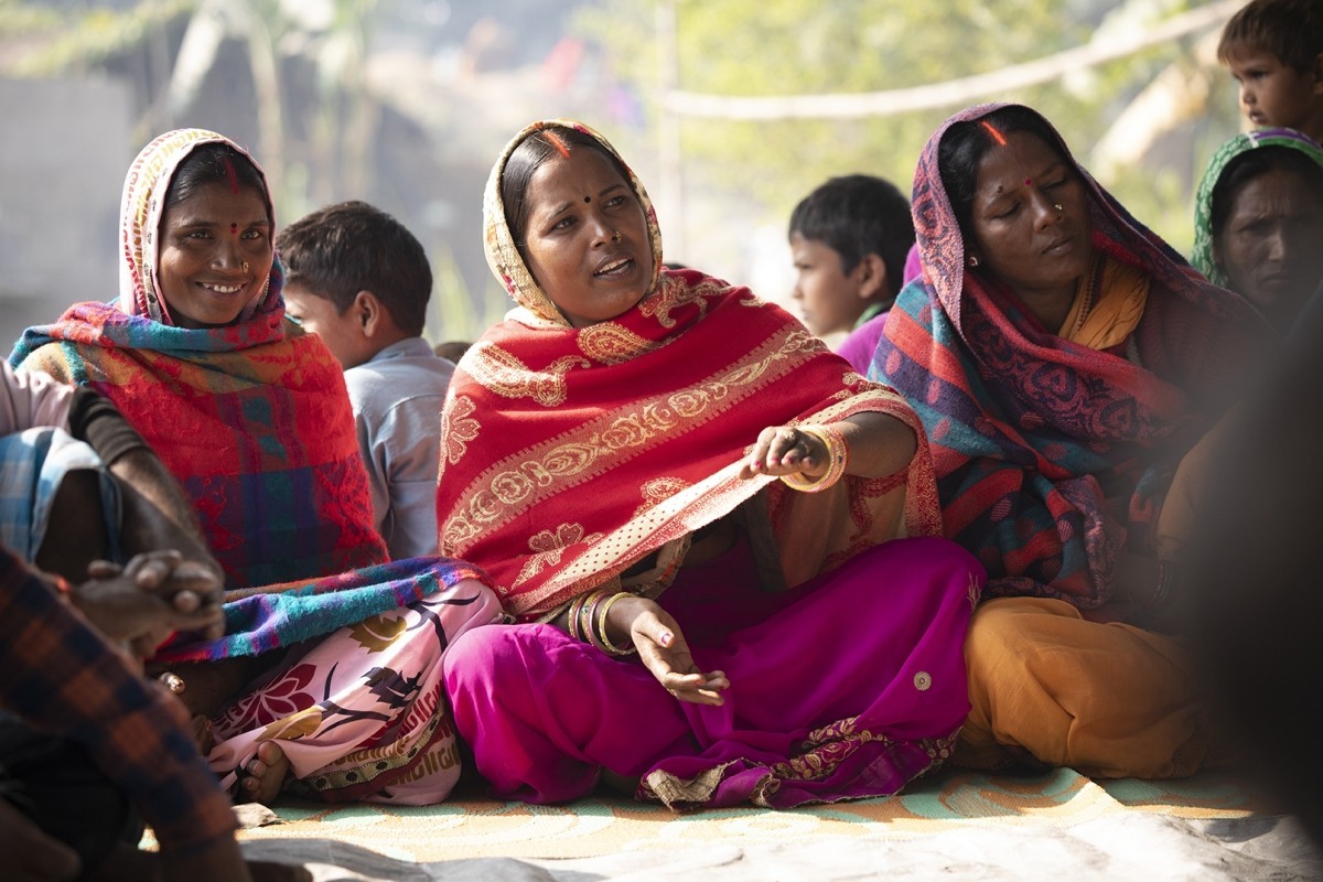 3 Indian women in brightly colored clothes sit next each other with the woman in the middle actively speaking