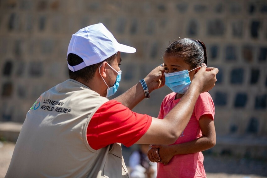 Man wearing white baseball hat, red t-shirt and tan vest with the LWR logo kneels to help a young girl in a pink t-shirt put on her mask