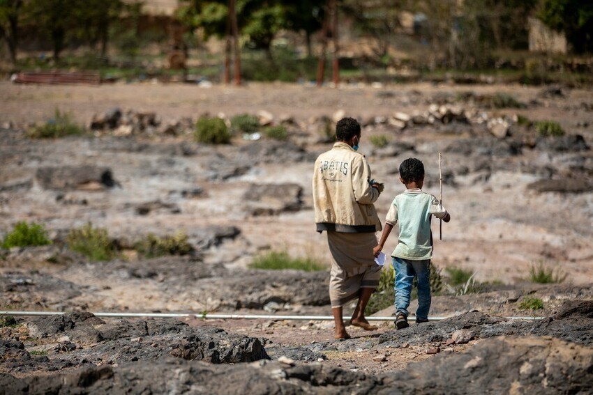 A young boy carrying a stick and a man without shoes walk across sand rocks and shrub away from the camera