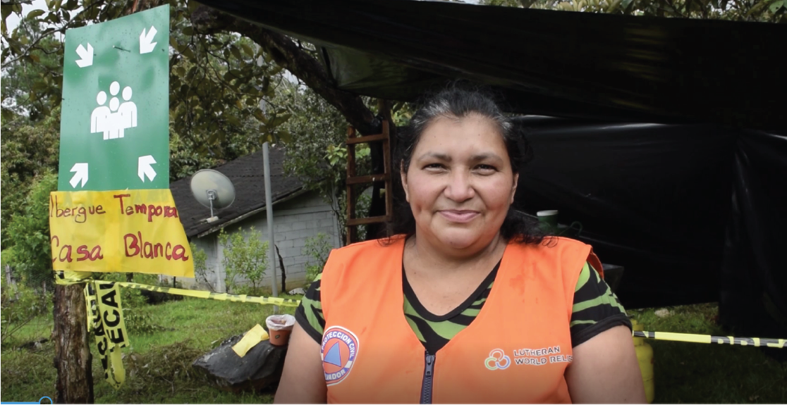 A woman in a bright orange safety vest with a Lutheran World Relief logo stands in front of a structure that is labelled temporary shelter in Spanish.