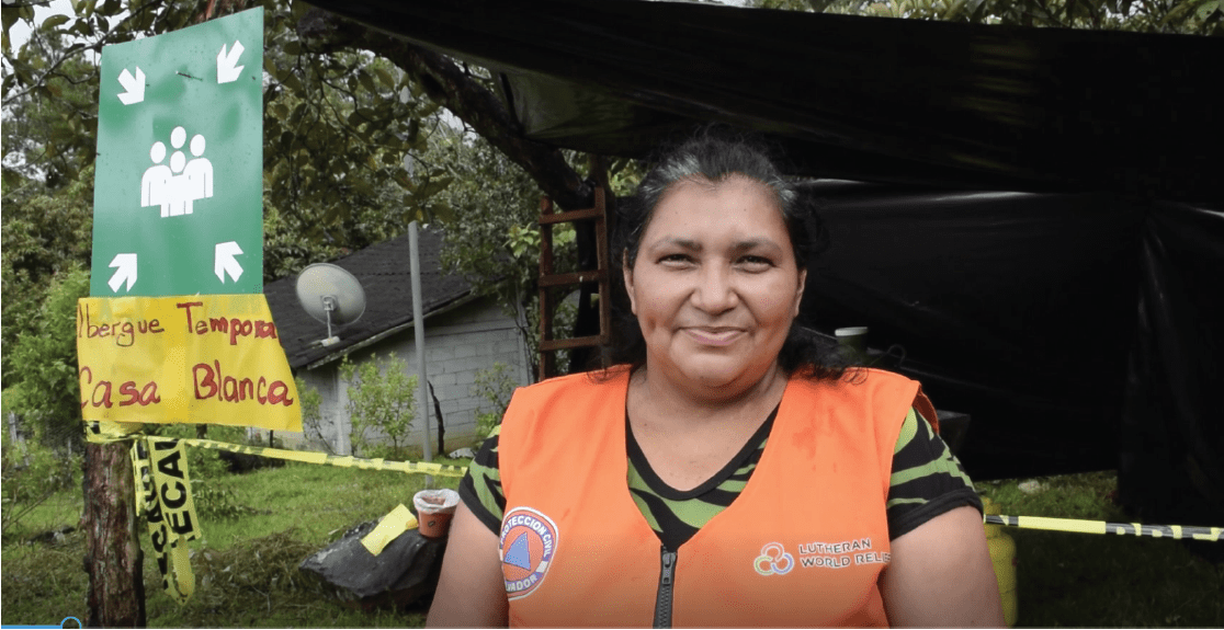 A woman in a bright orange safety vest with a Lutheran World Relief logo stands in front of a structure that is labelled temporary shelter in Spanish.