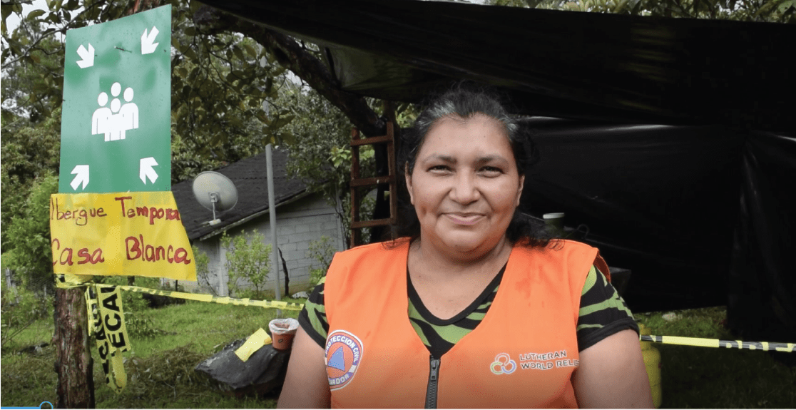 A woman in a bright orange safety vest with a Lutheran World Relief logo stands in front of a structure that is labelled temporary shelter in Spanish. 